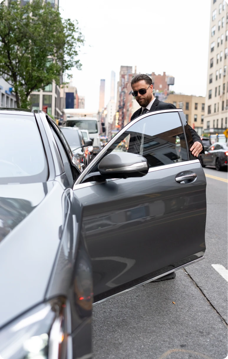 A security agent in a suit and sunglasses holding open the door of a gray luxury sedan on a city street.